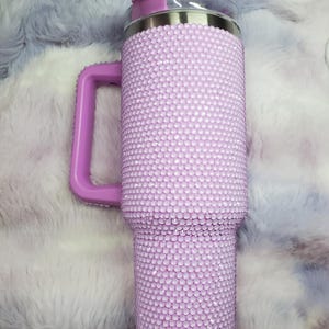 Pu&ograve; includere: Un bicchiere viola chiaro con manico, ricoperto di piccole e scintillanti strass. Il bicchiere ha un bordo argentato e un coperchio viola abbinato. Anche il manico &egrave; viola. Lo sfondo &egrave; un materiale soffice, viola chiaro e bianco.