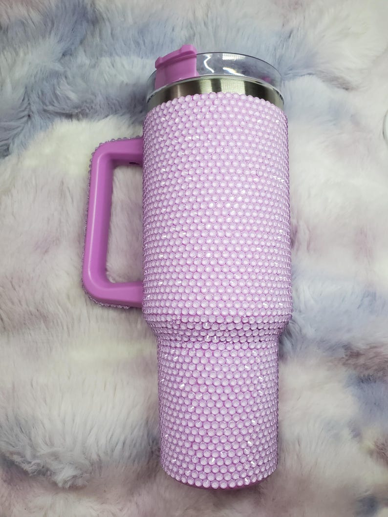 Pu&ograve; includere: Un bicchiere viola chiaro con manico, ricoperto di piccole e scintillanti strass. Il bicchiere ha un bordo argentato e un coperchio viola abbinato. Anche il manico &egrave; viola. Lo sfondo &egrave; un materiale soffice, viola chiaro e bianco.