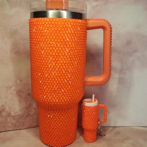 Pu&ograve; includere: Un bicchiere arancione tempestato di strass con manico e coperchio trasparente. Accanto c'&egrave; un bicchiere portachiavi pi&ugrave; piccolo abbinato. I bicchieri sono ricoperti da piccoli abbellimenti rotondi arancioni. Il manico e il coperchio sono di colore arancione pieno.