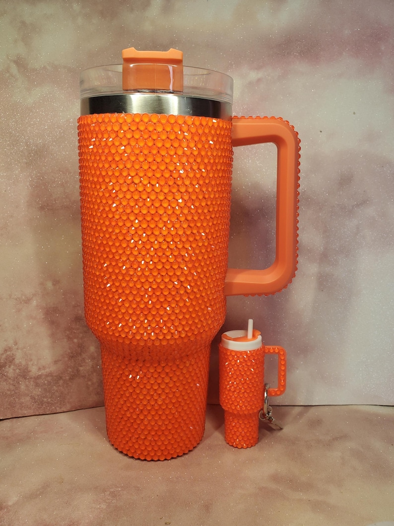 Pu&ograve; includere: Un bicchiere arancione tempestato di strass con manico e coperchio trasparente. Accanto c'&egrave; un bicchiere portachiavi pi&ugrave; piccolo abbinato. I bicchieri sono ricoperti da piccoli abbellimenti rotondi arancioni. Il manico e il coperchio sono di colore arancione pieno.