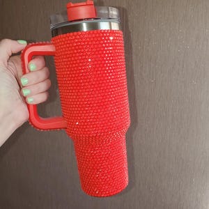 Pu&ograve; includere: Un bicchiere rosso ricoperto di strass scintillanti. Il bicchiere ha un manico rosso e un coperchio rosso con la parte superiore trasparente. Il bicchiere &egrave; tenuto da una mano con smalto verde chiaro.