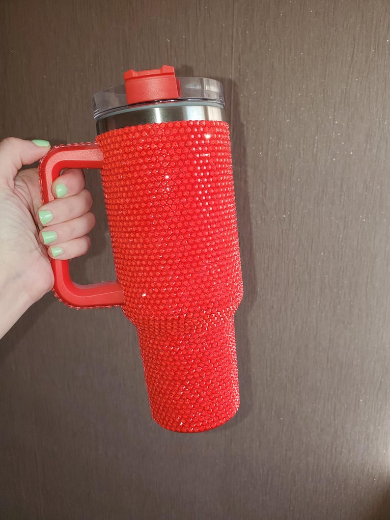 Pu&ograve; includere: Un bicchiere rosso ricoperto di strass scintillanti. Il bicchiere ha un manico rosso e un coperchio rosso con la parte superiore trasparente. Il bicchiere &egrave; tenuto da una mano con smalto verde chiaro.