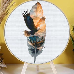 May include: A cross-stitch embroidery in a wooden hoop depicts a feather with an eagle in flight. The feather design features a sunset and a forest scene, with shades of orange, blue, and green. The embroidery is displayed on a wooden stand.