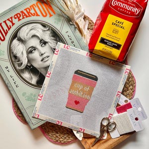 May include: A Dolly Parton book, a red bag of Community Coffee, a needlepoint canvas featuring a coffee cup with the text cup of ambition, small metal scissors, and two spools of thread. The canvas is marked with the Lagniappe Letter Co logo.