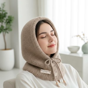 May include: A beige hooded scarf with a textured knit design. The scarf has a drawstring closure with wooden beads and a small label that reads "WoolSeasonCo". The scarf is worn over a white shirt.