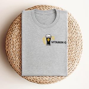 May include: A heather gray t-shirt with a crew neck. The shirt features a graphic of a beer glass with foam and a yellow design, along with the text "VITAMIN G" in black. The shirt is folded on a woven, circular surface.
