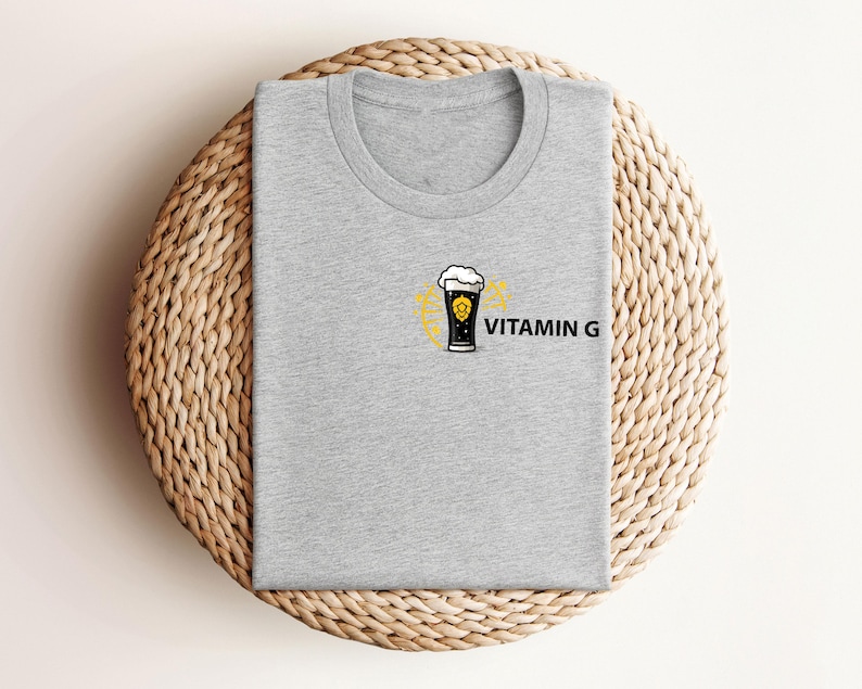 May include: A heather gray t-shirt with a crew neck. The shirt features a graphic of a beer glass with foam and a yellow design, along with the text "VITAMIN G" in black. The shirt is folded on a woven, circular surface.