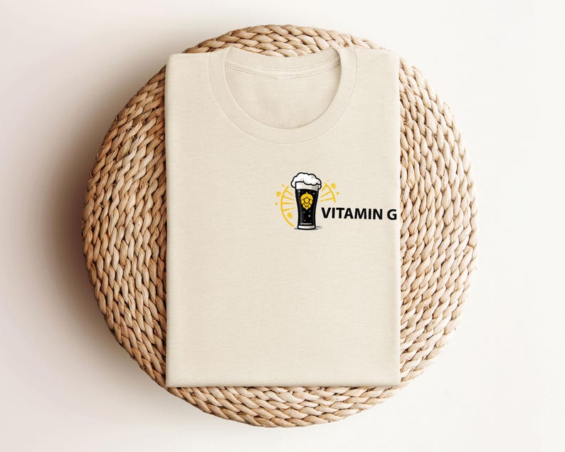 May include: A cream-colored t-shirt with a graphic of a beer glass with foam, surrounded by a yellow sunburst design. The text "VITAMIN G" is printed in black next to the graphic. The shirt is folded on a woven, circular surface.