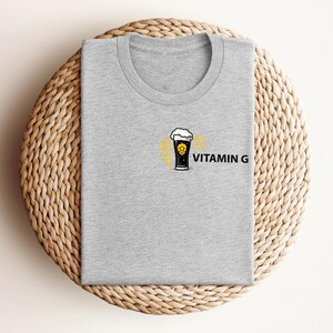 May include: A heather gray t-shirt with a crew neck. The shirt features a graphic of a beer glass with foam and a yellow design, along with the text "VITAMIN G" in black. The shirt is folded on a woven, circular surface.