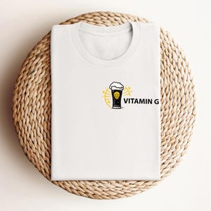 May include: White t-shirt with a small graphic of a beer glass and the words "VITAMIN G" in black. The beer glass is black with a yellow outline and a foamy top. The shirt is folded on a round, woven, light brown surface.