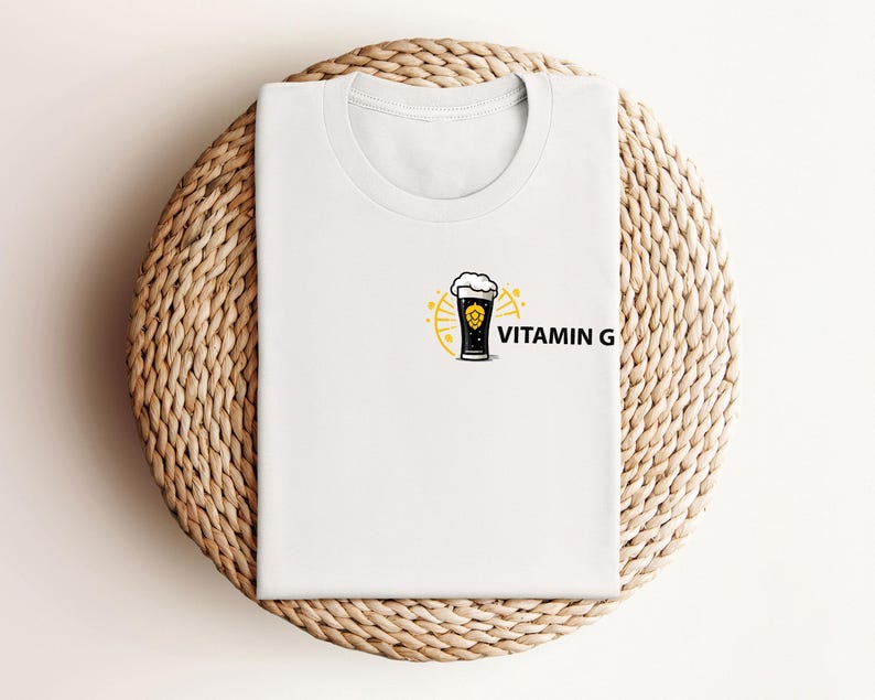 May include: White t-shirt with a small graphic of a beer glass and the words "VITAMIN G" in black. The beer glass is black with a yellow outline and a foamy top. The shirt is folded on a round, woven, light brown surface.