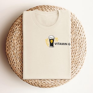 May include: A cream-colored t-shirt featuring a graphic of a beer glass with foam, surrounded by a yellow sunburst design. The text "VITAMIN G" is printed in black next to the beer glass. The shirt is folded on a woven, circular surface.