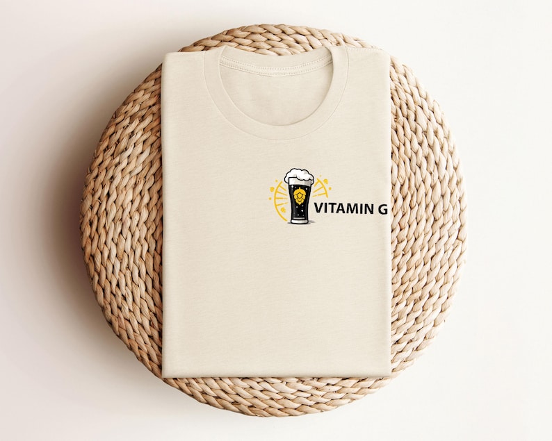 May include: A cream-colored t-shirt featuring a graphic of a beer glass with foam, surrounded by a yellow sunburst design. The text "VITAMIN G" is printed in black next to the beer glass. The shirt is folded on a woven, circular surface.