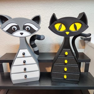 Op de afbeelding: Twee decoratieve sieradendoosjes: een grijze wasbeer met een gestreepte staart en een zwarte kat met gele ogen. Elke doos heeft een reeks kleine lades met knoppen. De wasbeerdos heeft witte lades en de kattenbox heeft zwarte lades.