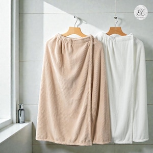 May include: Two soft bath wraps, one in beige and one in white, hang on wooden hangers. The wraps have a textured surface and a wrap-around design. Perfect for after a shower, they are a comfortable item.