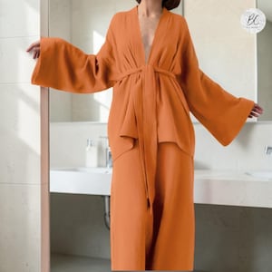 Può includere: Un completo arancione composto da una vestaglia e pantaloni abbinati. La vestaglia ha maniche ampie e una cintura annodata in vita. L'outfit è realizzato in un tessuto morbido e strutturato. Il set è esposto in un bagno.