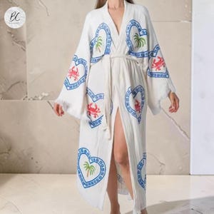 May include: White, long, open-front robe with a tie waist. The robe features blue heart-shaped designs with palm trees and red crab illustrations. The robe has wide sleeves and a split front, creating a flowing silhouette.