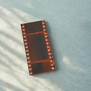 May include: A strip of film with a brown and orange hue, featuring sprocket holes along the edges. The film strip is rectangular and appears to be a photograph negative. The background is a light blue surface.