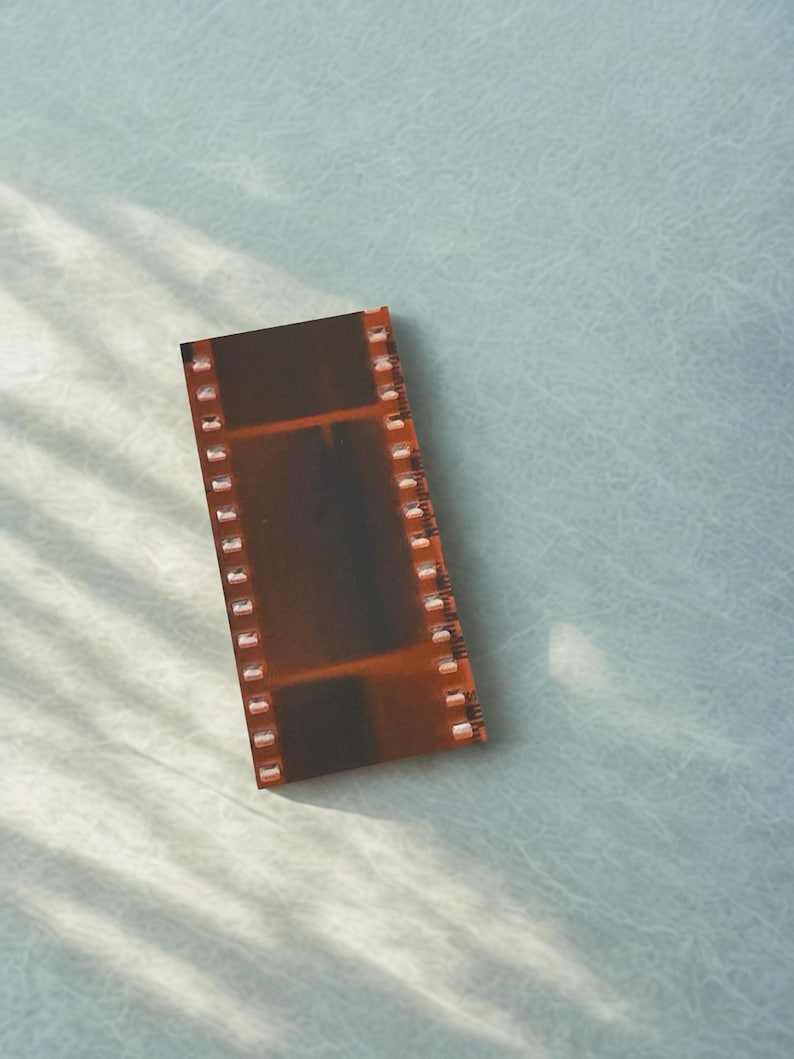 May include: A strip of film with a brown and orange hue, featuring sprocket holes along the edges. The film strip is rectangular and appears to be a photograph negative. The background is a light blue surface.