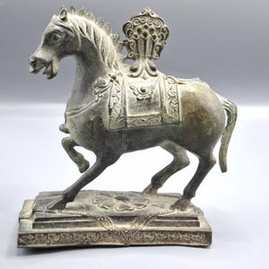 Könnte beinhalten: Bronzefarbene Pferdestatue mit detailliertem Sattel und dekorativem Kopfschmuck. Das Pferd ist im Schritt auf einem rechteckigen Sockel mit floralen Mustern dargestellt. Die Statue hat ein verwittertes Aussehen.