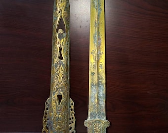 Op de afbeelding: Een antiek bronzen zwaard en schede. Het zwaard heeft een lang, slank lemmet met geëtste markeringen. De schede is ingewikkeld versierd met uitgesneden ontwerpen. Beide stukken vertonen tekenen van ouderdom en slijtage, met een groenachtige patina.