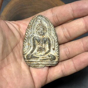 May include: A small, antique-looking Buddha amulet. The amulet is a dark, weathered color with intricate details of the seated Buddha figure. The amulet is oval-shaped and appears to be made of metal.