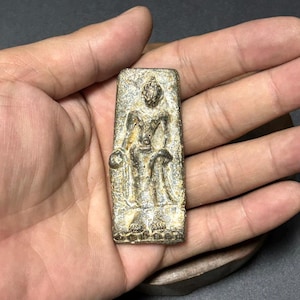 Puede incluir: Un amuleto metálico rectangular de aspecto envejecido, que representa una figura de pie en relieve. La figura está intrincadamente detallada, y el amuleto tiene un aspecto desgastado y antiguo. El color es un oro apagado con acentos más oscuros.