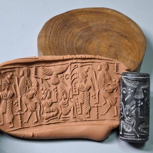 Puede incluir: Exhibición de artefactos antiguos con un relieve de terracota con tallas detalladas de figuras y animales. También hay un sello cilíndrico negro con tallas blancas, junto con un fondo de rodaja de madera. El relieve de terracota es de color marrón rojizo.