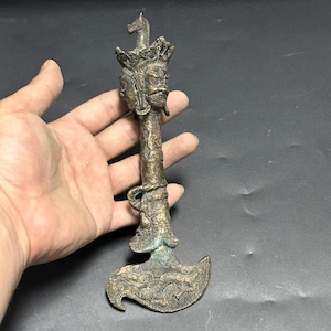 May include: A bronze-colored, ornate axe-shaped sculpture. The handle features detailed carvings and a figure with multiple faces and a horse head on top. The axe head has a dragon design.