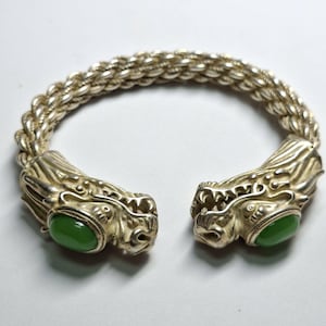 Puede incluir: Pulsera plateada con terminales de cabeza de dragón y detalles de piedra verde ovalada. La pulsera tiene un diseño de cadena trenzada. Las cabezas de dragón están detalladas con escamas y rasgos faciales.