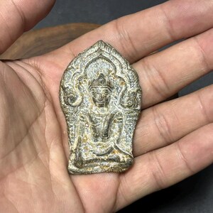 Puede incluir: Un pequeño amuleto de Buda de aspecto antiguo. El amuleto es de color gris claro con detalles intrincados, incluyendo una figura de Buda sentado con una corona. El amuleto se sostiene en una mano, mostrando su tamaño y textura. El diseño general sugiere un artículo religioso o espiritual.