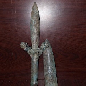 May include: An antique bronze dagger with a detailed handle and a separate sheath. The dagger has a pointed blade and intricate carvings on the handle and sheath. The bronze has a weathered, aged appearance.