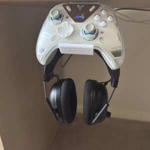 May include: A white gaming controller with blue accents and the word "APEXS" printed on it is mounted on a white holder. Black headphones are attached to the controller. The controller is hanging from a wooden shelf.