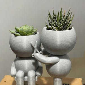 Puede incluir: Maceta gris impresa en 3D con forma de persona cortándose el pelo. Una maceta contiene una suculenta verde, la otra una planta espinosa. La persona está sentada y la otra sostiene unas tijeras. Las macetas están sobre una superficie marrón.