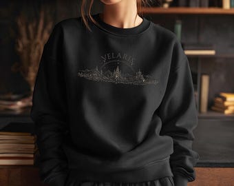 Puede incluir: Sudadera negra de cuello redondo con la palabra "VELARIS" arqueada sobre una ilustración dorada de un paisaje urbano. El diseño presenta edificios, montañas y un cielo estrellado. La sudadera es de material suave.