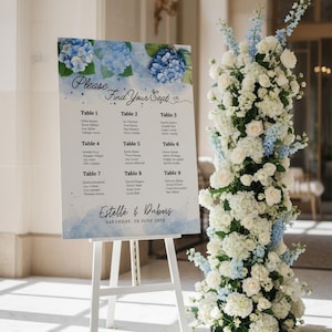 以下が含まれることがあります： 白いイーゼルには、青い水彩画の花柄と「Please Find Your Seat」の文字が入った結婚式の座席表が表示されています。チャートにはテーブル番号とゲストの名前が記載されています。右側には、白と青の花の背の高いフラワーアレンジメントがあります。