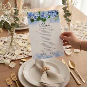 May include: A wedding menu with blue hydrangea floral design. The menu is held above a table setting with plates, gold cutlery, and a napkin ring. The menu reads "Menu" and includes the names "Estelle & Dubois" and the date "SATURDAY, 28 JUNE 2629."