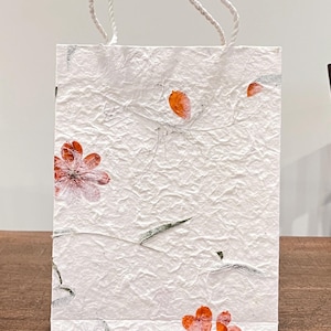 May include: A white paper gift bag with a textured surface and a twisted white rope handle. The bag is decorated with red and orange floral and leaf designs. The bag is sitting on a wooden surface.
