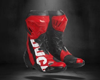 May include: Pair of red and black motorcycle boots with the word "DUCATI" in white lettering. The boots have a high-top design with protective elements and a mesh panel on the front. The boots are on a dark background.
