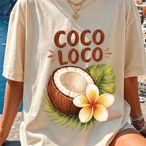 May include: Cream-colored t-shirt with the words "COCO LOCO" in brown, featuring a coconut, tropical flower, and green leaves. The shirt is a relaxed fit, perfect for a casual, beachy vibe.