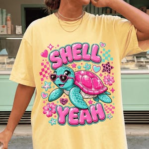 May include: Yellow t-shirt with a cartoon turtle wearing heart-shaped sunglasses and the words "SHELL YEAH" in pink and blue. The turtle has a pink shell and is surrounded by flowers and hearts.