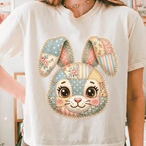 Pode incluir: Camiseta creme com um rosto de coelho patchwork colorido. O coelho tem olhos grandes e expressivos, um nariz rosa e orelhas longas. O design patchwork inclui padrões florais, de bolinhas e listrados, criando uma estética lúdica e charmosa.