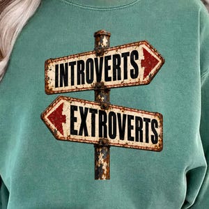 May include: A sage green crewneck sweatshirt featuring a graphic of a weathered, rusted metal signpost with two directional arrows. The top arrow points right and reads INTROVERTS in black text. The bottom arrow points left and reads EXTROVERTS.