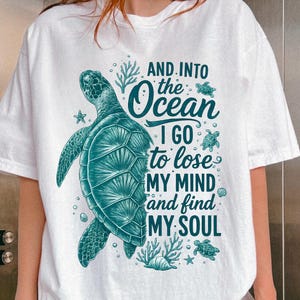 May include: A white short-sleeve t-shirt featuring a teal graphic of a sea turtle surrounded by small sea creatures and coral. The text reads: AND INTO THE OCEAN I GO TO LOSE MY MIND AND FIND MY SOUL. The design is centered on the front of the shirt.