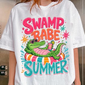 May include: A white t-shirt featuring a graphic of a green alligator wearing heart-shaped sunglasses, lounging on a striped pool float while holding a drink. The text reads SWAMP BABE SUMMER in bright pink, orange, and teal lettering with star accents.