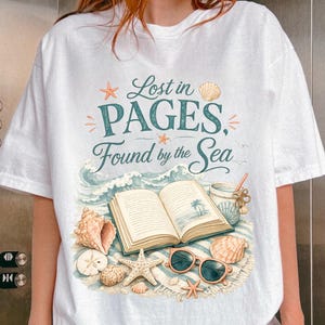 May include: A white t-shirt featuring a graphic print with the text Lost in PAGES, Found by the Sea. The design includes an open book, seashells, a starfish, a sand dollar, and sunglasses resting on a sandy beach with ocean waves.