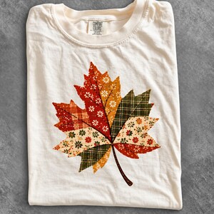 May include: A cream-coloured t-shirt featuring a large, central graphic of a maple leaf composed of various patchwork-style fabric patterns in shades of red, orange, green, and white, with a Comfort Colors brand label visible at the neckline.