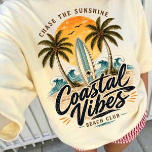 May include: Cream-colored t-shirt with a beach-themed graphic. The design features a sunset, palm trees, a surfboard, and ocean waves. Text includes "Chase the Sunshine" and "Coastal Vibes Beach Club."