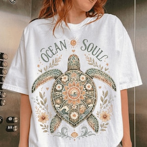 May include: White t-shirt featuring a detailed sea turtle design with the words "Ocean Soul". The turtle has a floral pattern on its shell, surrounded by flowers and leaves. The design incorporates shades of green, brown, and orange.