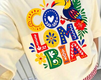 May include: A cream-colored t-shirt featuring a colorful graphic that spells out COLOMBIA. The design includes a toucan, a red hibiscus flower, green leaves, and various decorative patterns in yellow, blue, and red surrounding the letters.